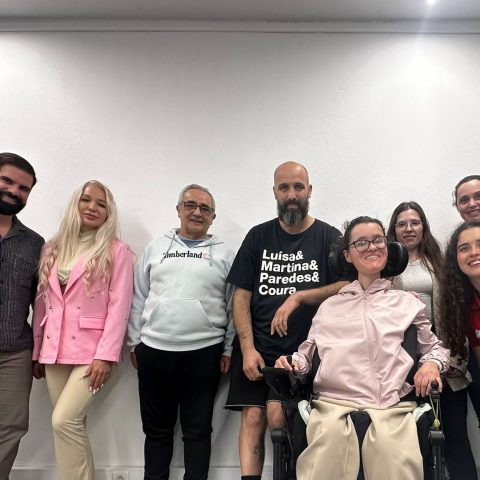 Um grupo de sete pessoas em pé e uma pessoa em cadeira de rodas, alinhadas lado a lado para uma foto em ambiente interior. O fundo é uma parede branca simples, com teto claro e luzes embutidas.