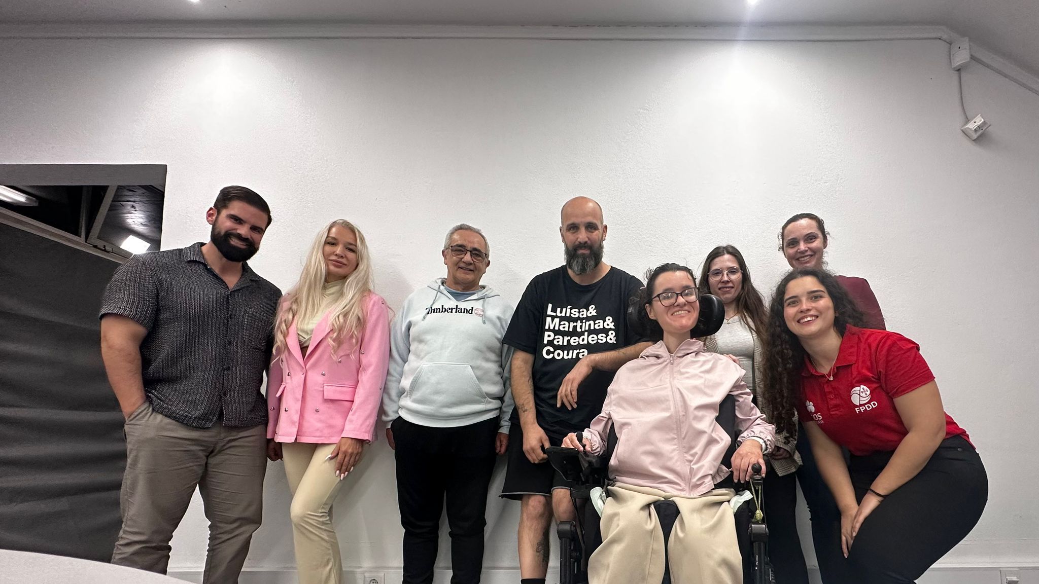 Um grupo de sete pessoas em pé e uma pessoa em cadeira de rodas, alinhadas lado a lado para uma foto em ambiente interior. O fundo é uma parede branca simples, com teto claro e luzes embutidas.
