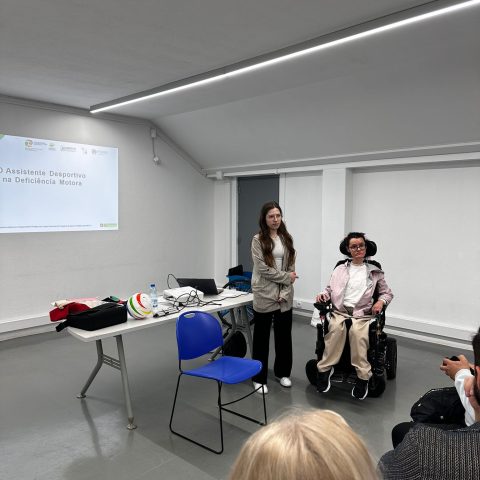 Sala de formação com uma apresentação sobre desporto e deficiência. Uma pessoa em cadeira de rodas elétrica à direita, posicionada de frente para o público. Ao lado, mais ao centro, está uma mulher de pé. À esquerda encontra‑se uma mesa branca retangular. Sobre a mesa há um computador portátil aberto, uma garrafa de água e uma bola multicolorida de plástico À frente da mesa há uma cadeira azul simples de plástico, sem ninguém sentado. No fundo, projetado na parede branca, há um slide com fundo claro. O título no slide diz: “Assistente Desportivo na Deficiência Motora”