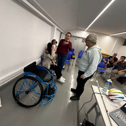 Sala ampla com cadeiras azuis e uma cadeira de rodas desportiva azul. À esquerda, uma mulher, está sentada numa cadeira azul, com a mão junto ao rosto. Ao lado dela, mais ao fundo, outra mulher de óculos, está sentada e observa a cena. No centro, uma mulher está de pé, com uma mão sobre o peito, em atitude de explicação ou conversa. À frente dela está um homem, virado para a mulher. No fundo, várias cadeiras azuis estão vazias e, à direita, três homens estão sentados, voltados para o grupo principal, parecendo ouvir;