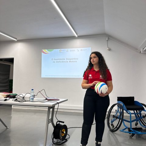 Mulher jovem de pé numa sala, em frente a um ecrã de projeção. Ela tem uma camisola polo vermelha da FPDD calças pretas justas e sapatilhas desportivas. Segura uma bola branca com vários segmentos coloridos em verde, rosa, azul e amarelo fluorescente. Atrás dela, projetado na parede branca, está um slide com o título: “O Assistente Desportivo na Deficiência Motora”. À esquerda, há uma mesa branca com tampo retangular. Em cima da mesa estão um computador portátil aberto, uma garrafa de água. À direita da mulher está uma cadeira de rodas desportiva azul.
