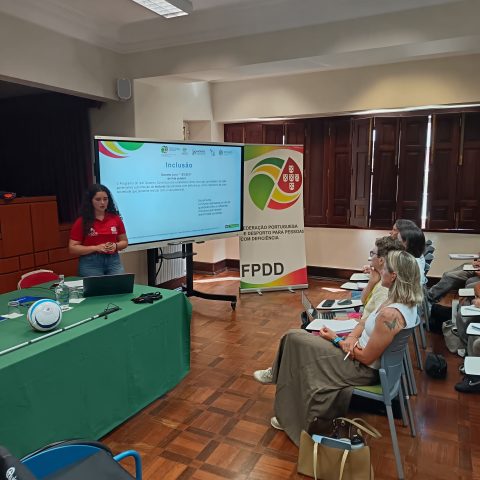 Sala de formação com uma apresentação sobre inclusão. Na imagem aparece uma mulher de pele clara e cabelo encaracolado castanho-escuro, com camisola vermelha e calças de ganga, em pé à esquerda, junto de uma mesa retangular coberta com toalha verde. Sobre a mesa há um computador portátil aberto, um bastão branco de mobilidade, uma bola branca com detalhes azuis, uma garrafa de água e alguns pequenos objetos. À frente da mulher, ligeiramente à direita, há um ecrã grande na horizontal com um slide projetado com o título “Inclusão” em letras azuis. À direita do ecrã encontra se um roll up vertical com o lógotipo da FPDD. Do lado direito da imagem estão várias pessoas sentadas em cadeiras individuais com mesas pequenas presas ao lado, em posição de audiência. No canto inferior esquerdo da imagem aparece parcialmente uma cadeira de rodas desportiva azul.