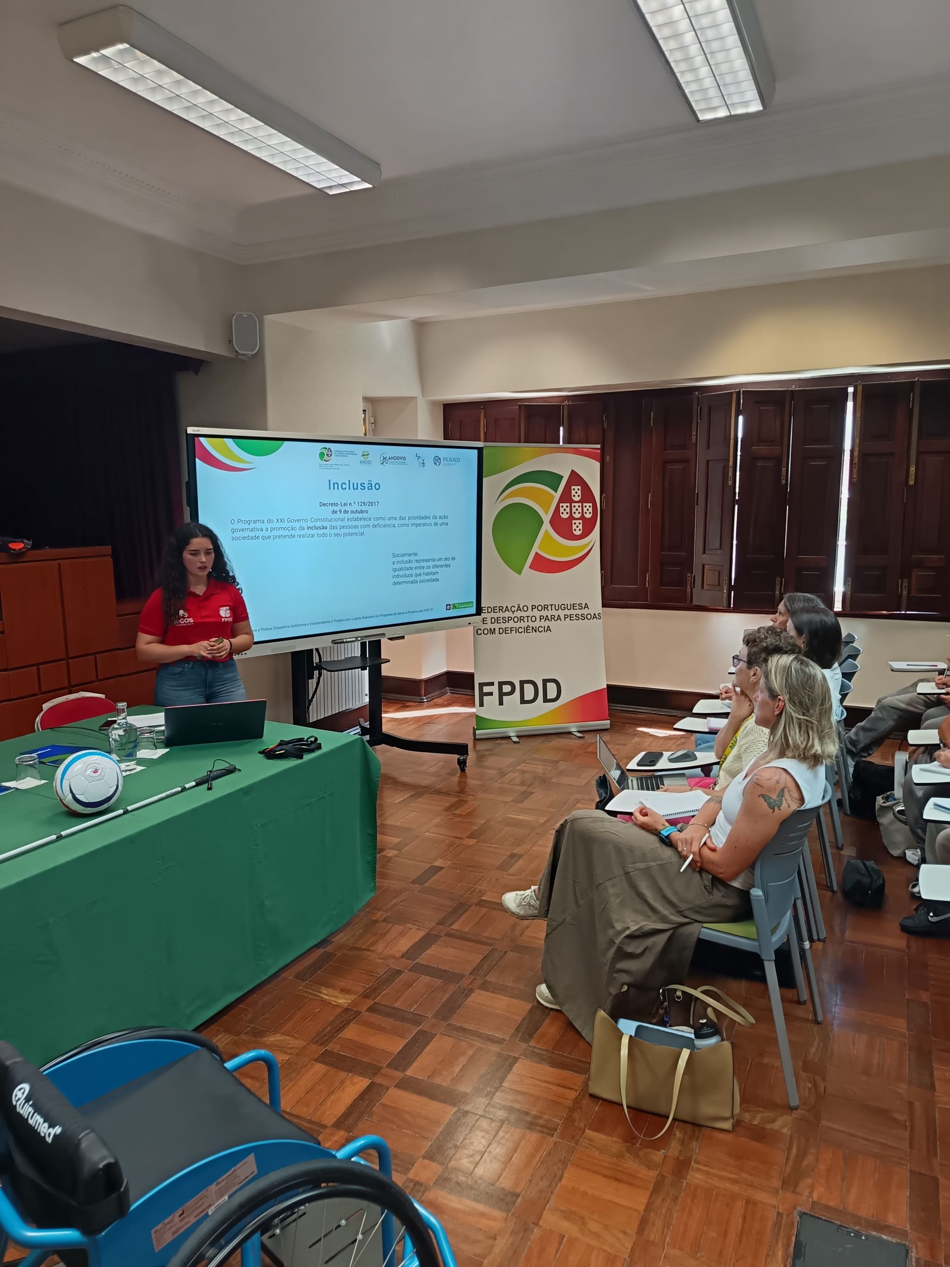 Sala de formação com uma apresentação sobre inclusão. Na imagem aparece uma mulher de pele clara e cabelo encaracolado castanho-escuro, com camisola vermelha e calças de ganga, em pé à esquerda, junto de uma mesa retangular coberta com toalha verde. Sobre a mesa há um computador portátil aberto, um bastão branco de mobilidade, uma bola branca com detalhes azuis, uma garrafa de água e alguns pequenos objetos. À frente da mulher, ligeiramente à direita, há um ecrã grande na horizontal com um slide projetado com o título “Inclusão” em letras azuis. À direita do ecrã encontra se um roll up vertical com o lógotipo da FPDD. Do lado direito da imagem estão várias pessoas sentadas em cadeiras individuais com mesas pequenas presas ao lado, em posição de audiência. No canto inferior esquerdo da imagem aparece parcialmente uma cadeira de rodas desportiva azul.