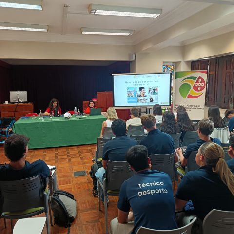 Sala de formação com pessoas sentadas a assistir a uma apresentação. Fileiras de cadeiras ocupadas por jovens, muitos a usar camisolas azuis-escuro com as palavras “Técnico Desporto” impressas nas costas em branco. À frente, uma mesa comprida coberta com toalha verde, com duas mulheres sentadas atrás de portáteis e vários objetos em cima da mesa, como garrafas de água, copos e papéis. Por trás da mesa, um ecrã de projeção mostra um slide de apresentação; À direita do ecrã encontra se um roll up vertical com o logotipo da FPDD.