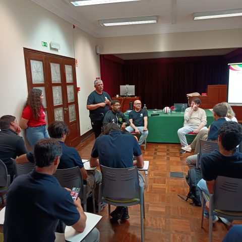 Sala de formação com várias pessoas sentadas a assistir à formação. Na frente da sala há uma mesa retangular coberta com toalha verde, onde estão quatro homens. Sobre a mesa há um monitor de computador, um teclado, uma garrafa de água, um copo e uma bola de voleibol branca com faixas vermelhas e verdes. Atrás da mesa existe um palco baixo com cortina escura e um computador de secretária ao fundo. À esquerda, perto de uma porta está um homem de cabelo grisalho; ao lado dele, mais perto da porta, está uma mulher de cabelo comprido encaracolado castanho, pele clara, com T-shirt vermelha, encostada à parede. Em primeiro plano e ao centro, vários participantes. À direita, perto da frente da sala, há um ecrã de projeção onde se vê parcialmente um slide com fundo branco e uma faixa colorida no canto superior direito.