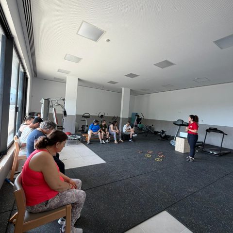 Sala de ginástica com um grupo de pessoas na Ação de Sensibilização. À esquerda, quatro pessoas, e à direita, uma formadora a usar polo vermelho a falar com o grupo.