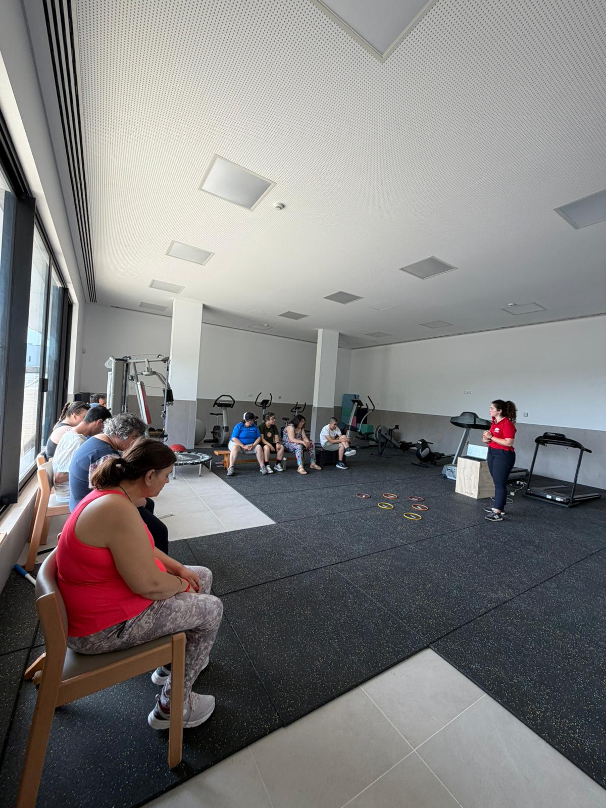 Sala de ginástica com um grupo de pessoas na Ação de Sensibilização. À esquerda, quatro pessoas, e à direita, uma formadora a usar polo vermelho a falar com o grupo.