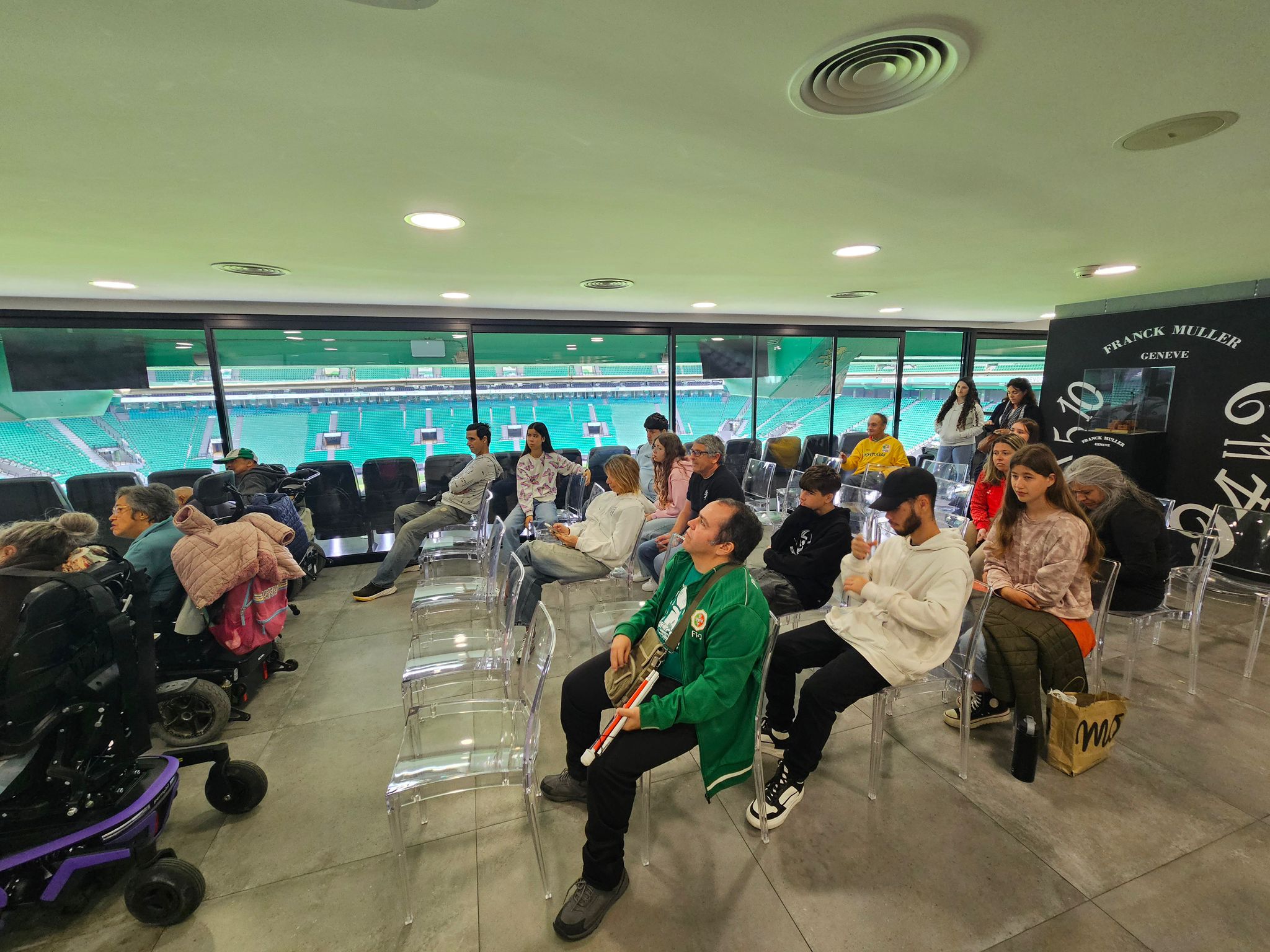 Sala de eventos com várias pessoas sentadas em cadeiras transparentes, diante de uma grande janela que dá para um estádio de futebol de arquibancadas verdes. A sala tem piso de tom cinza e teto branco com luminárias redondas embutidas e saídas de ventilação circulares. As cadeiras estão organizadas em fileiras; algumas estão vazias e outras ocupadas por adultos e alguns jovens.