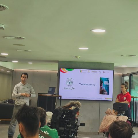 A imagem mostra uma apresentação em uma sala. Na frente, à esquerda, está um homem a falar para o público. Ao centro, há um ecrã grande com um slide projetado. No slide aparece o logótipo da Fundação Sporting Clube de Portugal e a palavra “Testemunhos. À direita do ecrã está uma mulher, em posição de atenção. À frente deles, estão pessoas sentadas, algumas em cadeiras de rodas, ouvindo a apresentação.