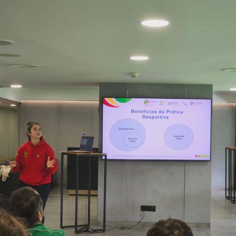 A foto mostra uma mulher a fazer uma apresentação numa sala. Ela está à esquerda, de pé. Tem os braços ligeiramente abertos, como se estivesse a explicar algo ao grupo à sua frente. Ao centro está um ecrã grande com um slide. O título diz “Benefícios da Prática Desportiva”. Abaixo há dois círculos: Círculo da esquerda, maior, com o texto “Atividade Física” e, dentro dele, um círculo menor com “Exercício Físico”. Círculo da direita, separado, com o texto “Inatividade Física”. Alguns participantes aparecem de costas, sentados em cadeiras transparentes.
