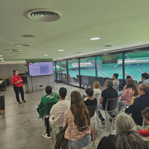 A imagem mostra a mesma sessão formativa numa sala com vista para o estádio do Sporting Clube de Portugal. À esquerda, à frente, está a apresentadora de hoodie vermelho e calças pretas, a falar para o grupo, perto do ecrã grande onde continua o tema “Benefícios da Prática Desportiva”. O slide atual tem uma tabela de texto, difícil de ler em detalhe, com vários pontos listados. À direita e ao fundo, várias pessoas estão sentadas em cadeiras transparentes, viradas para a apresentadora e para o ecrã.
