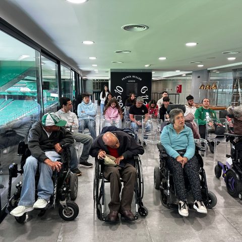 A foto mostra um grupo grande de pessoas numa sala com vista para o estádio. Na primeira fila, há quatro pessoas em cadeiras de rodas manuais ou elétricas. Atrás deles há várias filas de cadeiras transparentes com mais pessoas sentadas, entre jovens e adultos.