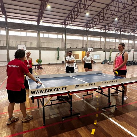 Participantes do Bootcamp 2026 assistem a uma demonstração de Polybat num pavilhão desportivo, enquanto uma formadora explica a modalidade adaptada à volta de uma mesa de jogo.