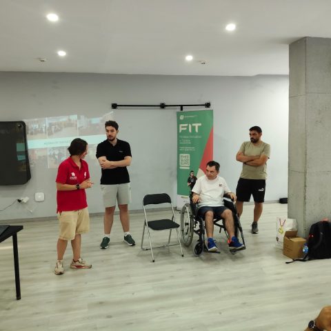 Momento de demonstração prática sobre exercício inclusivo, com um participante em cadeira de rodas e vários formandos reunidos numa sala de formação.