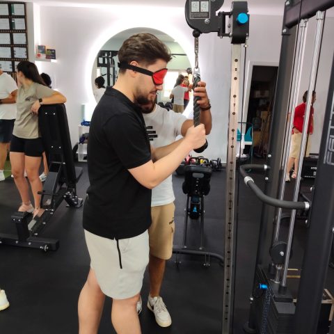 Participante realiza um exercício numa máquina de musculação com venda nos olhos, acompanhado por um formador, durante a ação de sensibilização.
