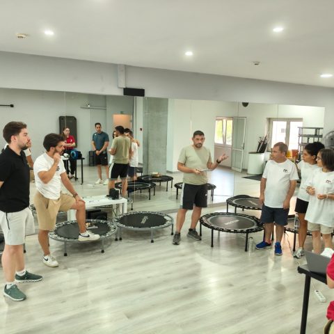 Grupo de participantes reúne-se em redor de minitrampolins enquanto um formador explica uma atividade prática de fitness inclusivo.