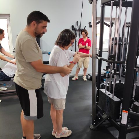 Formador acompanha uma participante durante a execução de um exercício numa máquina de musculação adaptada, enquanto outros participantes observam.