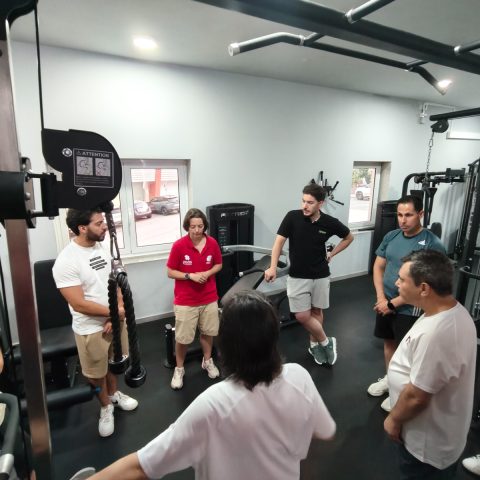 Grupo de participantes e formadores reúne-se numa sala de musculação para discutir estratégias de adaptação de exercícios a pessoas com deficiência.