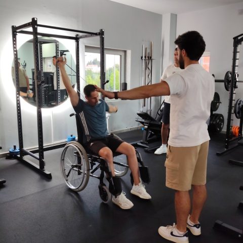 Participante em cadeira de rodas realiza um exercício de fortalecimento dos membros superiores com elástico, orientado por um formador.