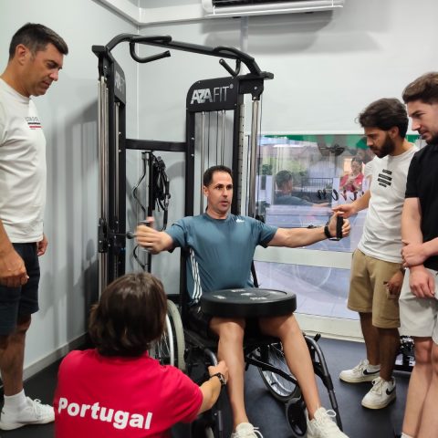 Participante em cadeira de rodas executa um exercício de peitoral numa máquina de musculação adaptada, acompanhado por formadores.