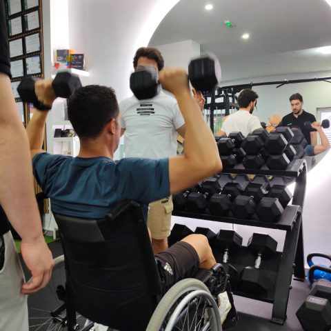 Participante em cadeira de rodas realiza um exercício com halteres sob supervisão de um formador, durante uma sessão prática de fitness inclusivo.